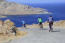 Drei Radfahrer radeln auf einer Straße auf der griechischen Insel Patmos entlang - im Hintergrund ist das tiefblaue Meer zu sehen