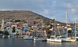 Blick zum Hafen mit Segel- und Fischerbooten sowie der Promenade mit den bunten Häusern in Symi auf der gleichnamigen Insel in der Südlichen Ägäis in Griechenland