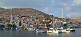 Blick zum Hafen mit Segel- und Fischerbooten sowie der Promenade mit den bunten Häusern in Symi auf der gleichnamigen Insel in der Südlichen Ägäis in Griechenland