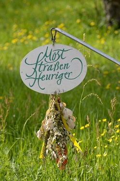 Ein uriges Heurigenschild in Österreich vor einer gelb blühenden Wiese.