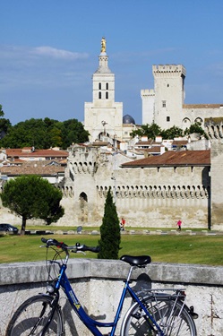 Ein Fahrrad lehnt vor dem Papstpalast von Avignon und der Kathedrale Notred-Dame-des-Doms an einer Mauer.