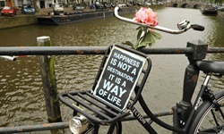 Ein altes Fahrrad mit einer Blume auf dem Lenker und einem Schild mit der Aufschrift "HAPPINIES IST NOT A DESTINATION, IT IS A WAY OF LIFE" auf einer Brücke über einen Fluss in Holland