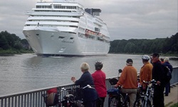 Eine Radlergruppe blickt auf den Nord-Ostsee-Kanal, in den ein großes Kreuzfahrtschiff einläuft