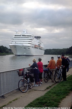Eine Radlergruppe blickt auf den Nord-Ostsee-Kanal, in den ein großes Kreuzfahrtschiff einläuft