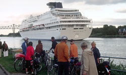 Eine Gruppe Radler blickt auf den Nord-Ostsee-Kanal, auf dem ein Kreuzfahrtschiff fährt