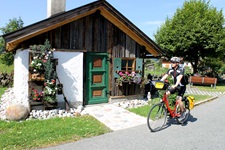 Ein Radler fährt auf einem asphaltierten Radweg an einem hübschen Holzhäuschen vorbei.