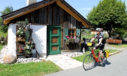 Ein Radler fährt auf einem asphaltierten Radweg an einem hübschen Holzhäuschen vorbei.