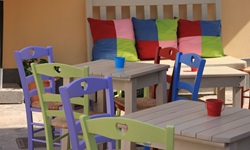 Holztische mit grünen und blauen Stühlen im Vordergrund. Hinten an einer Wand ist eine Holzbank mit bunten Kiseen zu sehen