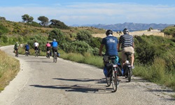 Eine Gruppe Radfahrer fährt auf einer Straße auf der Ionischen Insel Korfu entlang - im Hintergrund ist das Meer ganz leicht zu erkennen