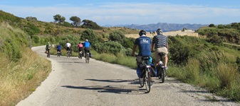 Eine Gruppe Radfahrer fährt auf einer Straße auf der Ionischen Insel Korfu entlang - im Hintergrund ist das Meer ganz leicht zu erkennen