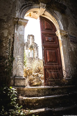 Ein halb offen stehendes Holzportal gibt den Blick auf eine steinerne Kirchenruine frei.