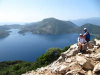Zwei Personen mit Radhelmen sitzen auf einem Berg, im Hintergrund ist das Meer und eine Ionische Insel zu erkennen