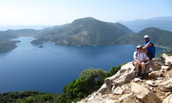 Zwei Personen mit Radhelmen sitzen auf einem Berg, im Hintergrund ist das Meer und eine Ionische Insel zu erkennen