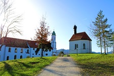 Ein von Kirche und Kapelle flankierter, weiß gekiester Radweg am Riegsee.