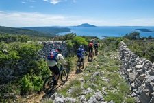 Vier Mountainbiker fahren auf einem von Steinmäuerchen eingerahmten Naturweg auf das tiefblaue Meer der Kvarner Bucht zu.
