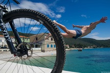 Nach erfolgreich absolvierter Tagestour wieder zurück im Hafen, gönnt sich ein Mountainbiker einen erfrischenden Sprung ins kühle Nass. Im Bildvordergrund ist das Vorderrad seines Mountainbikes zu erkennen.