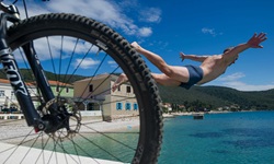 Nach erfolgreich absolvierter Tagestour wieder zurück im Hafen, gönnt sich ein Mountainbiker einen erfrischenden Sprung ins kühle Nass. Im Bildvordergrund ist das Vorderrad seines Mountainbikes zu erkennen.