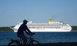 Ein Radfahrer fährt an der Ostseeküste entlang und im Hintergrund ist ein Krauzfahrtschiff auf dem Meer zu sehen