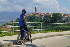 Ein Fahrradfahrer steht an einem Straßenrand auf der Insel Krk in Kroatien und blickt in die Kamera - hinter ihm ist die Gemeinde Vrbnik mit Kirchturm zu erkennen