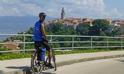 Ein Fahrradfahrer steht an einem Straßenrand auf der Insel Krk in Kroatien und blickt in die Kamera - hinter ihm ist die Gemeinde Vrbnik mit Kirchturm zu erkennen