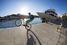 Ein Mountainbiker mit grüner Hose macht einen Wheelie an einer Hafenpromenade in der Kvarner Bucht in Kroatien
