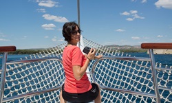 Eine Frau mit rotem T-Shirt und Sonnenbrille sitzt am Bug der Andela Lora, hält eine Kamera in der Hand und schaut in die Kamera