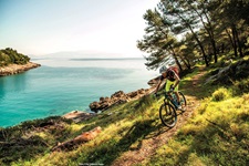 Ein Mountainbiker bei einer rasanten Abfahrt auf einem Naturweg, der parallel zum türkisblauen Meer der Kvarner Bucht verläuft.