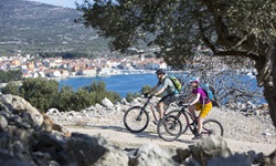 Ein Mountainbiker-Paar fährt auf einem schottrigen Weg auf der Insel Cres in der Kvarner Bucht von Kroatien entlang - Im Hintergrund ist eine Stadt und das angrenzende Meer zu sehen