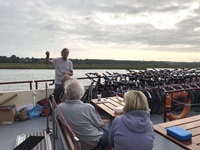 Ein Reiseleiter erläutert zwei Passagieren auf dem Sonnendeck den weiteren Ablauf des Tages. Neben ihm warten die aufgereihten Fahrräder auf ihren Einsatz.