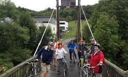Eine Radfahrergruppe in Lothringen schiebt ihre Räder auf einer hölzernen Brücke über den Fluss Sauer.