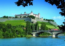 Ein Schloss auf einem Grasberg. Im Vordergrund sieht man den Main mit einer Brücke