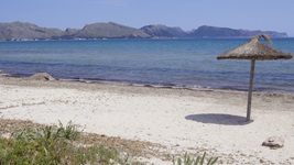 Blick auf einen Sandstrand mit einem Sonnenschirm aus Bast und Blick zum Gebirge auf Mallorca