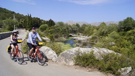 Zwei Radfahrer fahren auf einer asphaltierten Straße auf Mallorca an einem See vorbei, während im Hintergrund das imposante Gebirge zu sehen ist