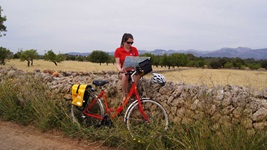 Radfahrerin sitzt auf einer Steinmauer und sieht sich auf einer Karte die Wegbeschreibung an, während sie ihr rotes Fahrrad vor der Mauer abgestellt hat