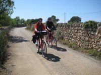 Zwei Radfahrer radeln über eine geschotterte Straße an einer Steinmauer auf Mallorca vorbei