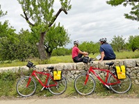 Zwei Radfahrer sitzen auf einer Steinmauer und blicken in die mallorquinische Landschaft, während ihre roten Fahrräder an der Steinmauer lehnen