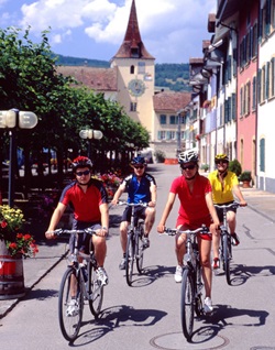 Vier Radfahrer radeln auf der Mittelland-Route durch einen Ort mit bunten Häuserfassaden. Im Hintergrund ist ein bemaltes Stadttor zu erkennen.