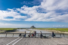 Vier Radler blicken von einer hölzernen Brücke zur Klosterinsel Mont St. Michel hinüber.