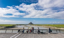 Vier Radler blicken von einer hölzernen Brücke zur Klosterinsel Mont St. Michel hinüber.