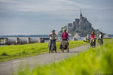 Eine Radlergruppe lässt auf einem von sattgrünem Gras gesäumten Radweg die Klosterinsel Mont St. Michel hinter sich.