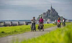 Eine Radlergruppe lässt auf einem von sattgrünem Gras gesäumten Radweg die Klosterinsel Mont St. Michel hinter sich.