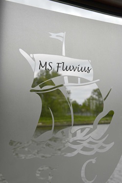 Detailbild eines Fensters der Fluvius mit Fensterbild, das ein Schiff mit der Aufschrift "MS Fluvius" zeigt