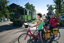 Die grüne Chiemsee Bahn passiert einen unbeschrankten Bahnübergang, an dem vier Radfahrer warten