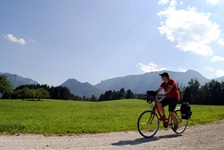 Ein Radfahrer radelt auf einem geschotterten Weg an Wiesen und Wälder vorbei - im Hintergrund die einzigartige Alpenkulisse