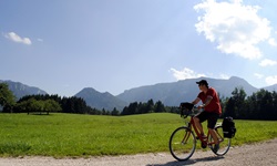 Ein Radfahrer radelt auf einem geschotterten Weg an Wiesen und Wälder vorbei - im Hintergrund die einzigartige Alpenkulisse