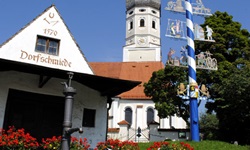 Zwei Räder stehen an einer Mauer hinter einem kleinen Dorfbrunnen angelehnt - hinter den Rädern ist ein Haus mit der Aufschrift "Dorfschmiede" und eine Kirche zu sehen