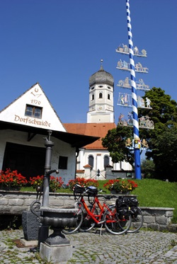 Zwei Räder stehen an einer Mauer hinter einem kleinen Dorfbrunnen angelehnt - hinter den Rädern ist ein Haus mit der Aufschrift "Dorfschmiede" und eine Kirche zu sehen