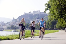Drei Radfahrer radeln entlang der Salzach - im Hintergrund ist die Festung Hohensalzburg zu sehen