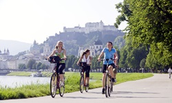 Drei Radfahrer radeln entlang der Salzach - im Hintergrund ist die Festung Hohensalzburg zu sehen