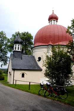 Eine große Kapelle mit einem runden Dach - davor sind am Holzgeländer zwei Räder abgestellt